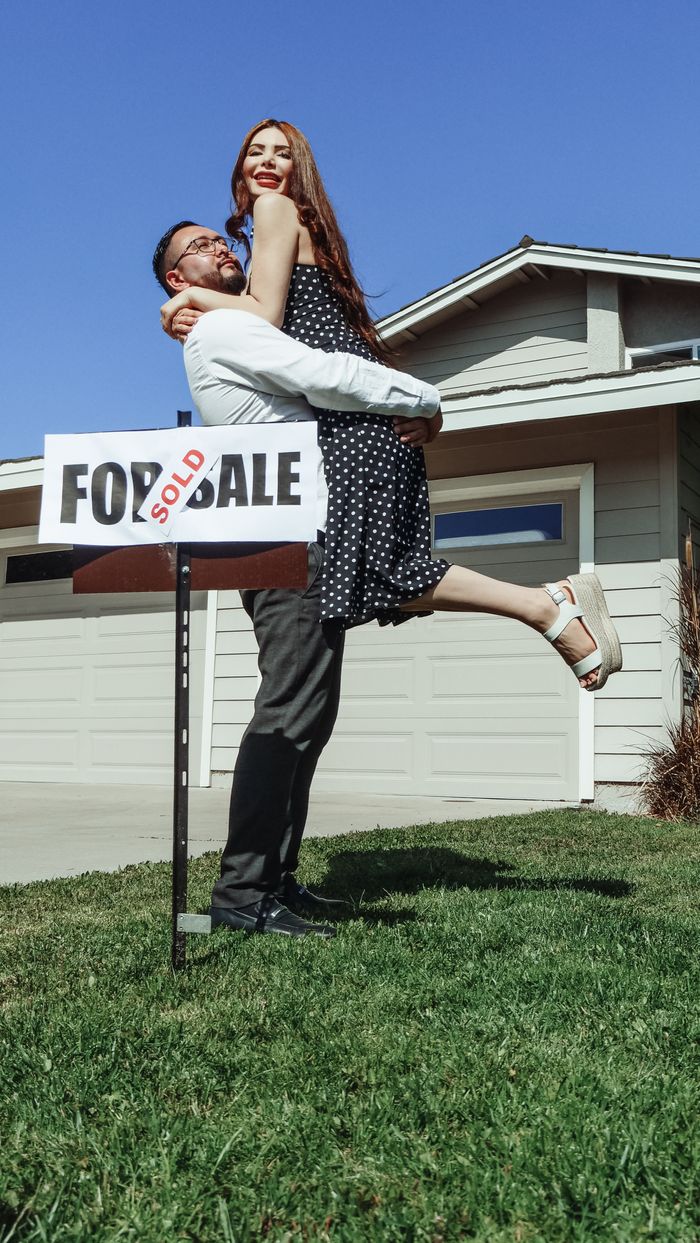 A joyful couple celebrates buying their new house with a 'Sold' sign.
