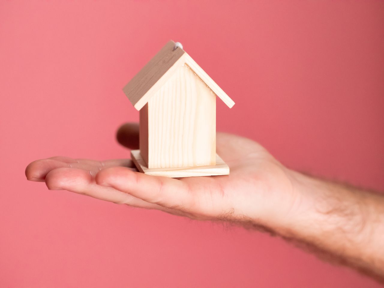 Wooden house model in hand symbolizing real estate concepts on pink background.
